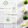 Set of cocktail-themed napkins with lime and bottle illustrations on a light surface with limes and lemon wedges.