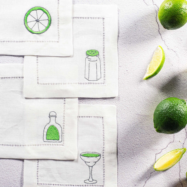 Set of cocktail-themed napkins with lime and bottle illustrations on a light surface with limes and lemon wedges.
