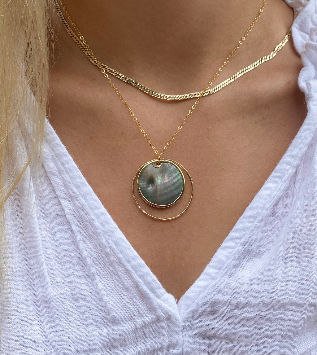 A close-up of a person wearing a layered gold necklace with a circular abalone shell pendant, paired with a white top.