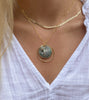 A close-up of a person wearing a layered gold necklace with a circular abalone shell pendant, paired with a white top.