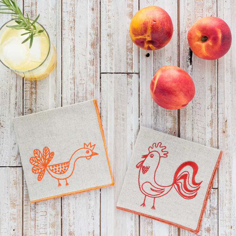Two cloth napkins with rooster designs on a wooden surface with peaches and a cocktail.
