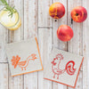 Two cloth napkins with rooster designs on a wooden surface with peaches and a cocktail.