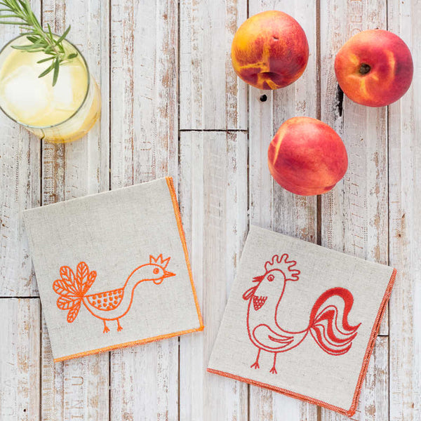 Two cloth napkins with rooster designs on a wooden surface with peaches and a cocktail.