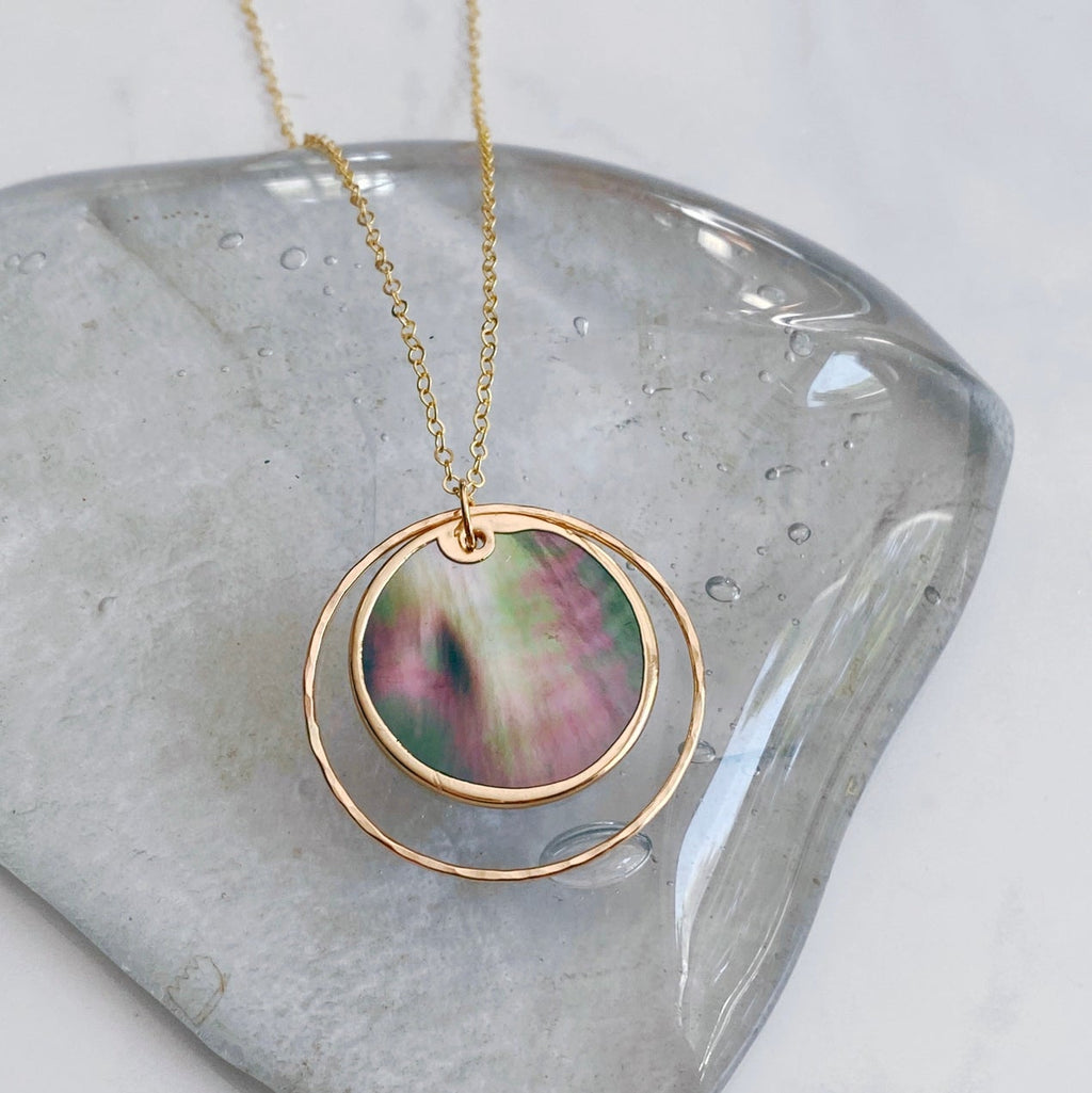 A gold necklace with a circular pendant featuring a colorful iridescent shell inlay, displayed on a smooth gray stone against a light marble background.