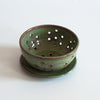 Medium ceramic bowl overtop a small saucer plate. The bowl has multiple groups of 7 holes divided around the surface and the bottom. A small amount of the original brown clay color pokes through the edges of the rims.