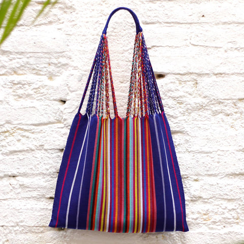 A colorful woven tote bag with a blue base and vertical stripes in red, orange, yellow, green, and white, hanging against a white brick wall.
