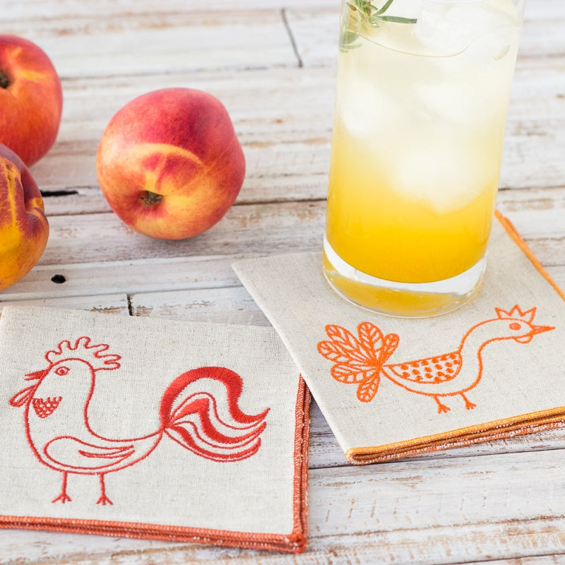Two embroidered napkins with rooster designs on a wooden surface with peaches and a glass of juice.