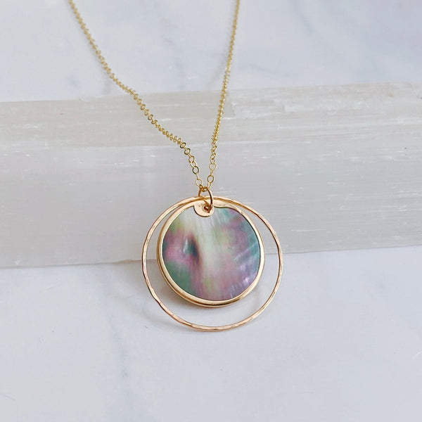 A gold necklace with a circular pendant featuring an iridescent abalone shell inlay, displayed on a light marble surface.