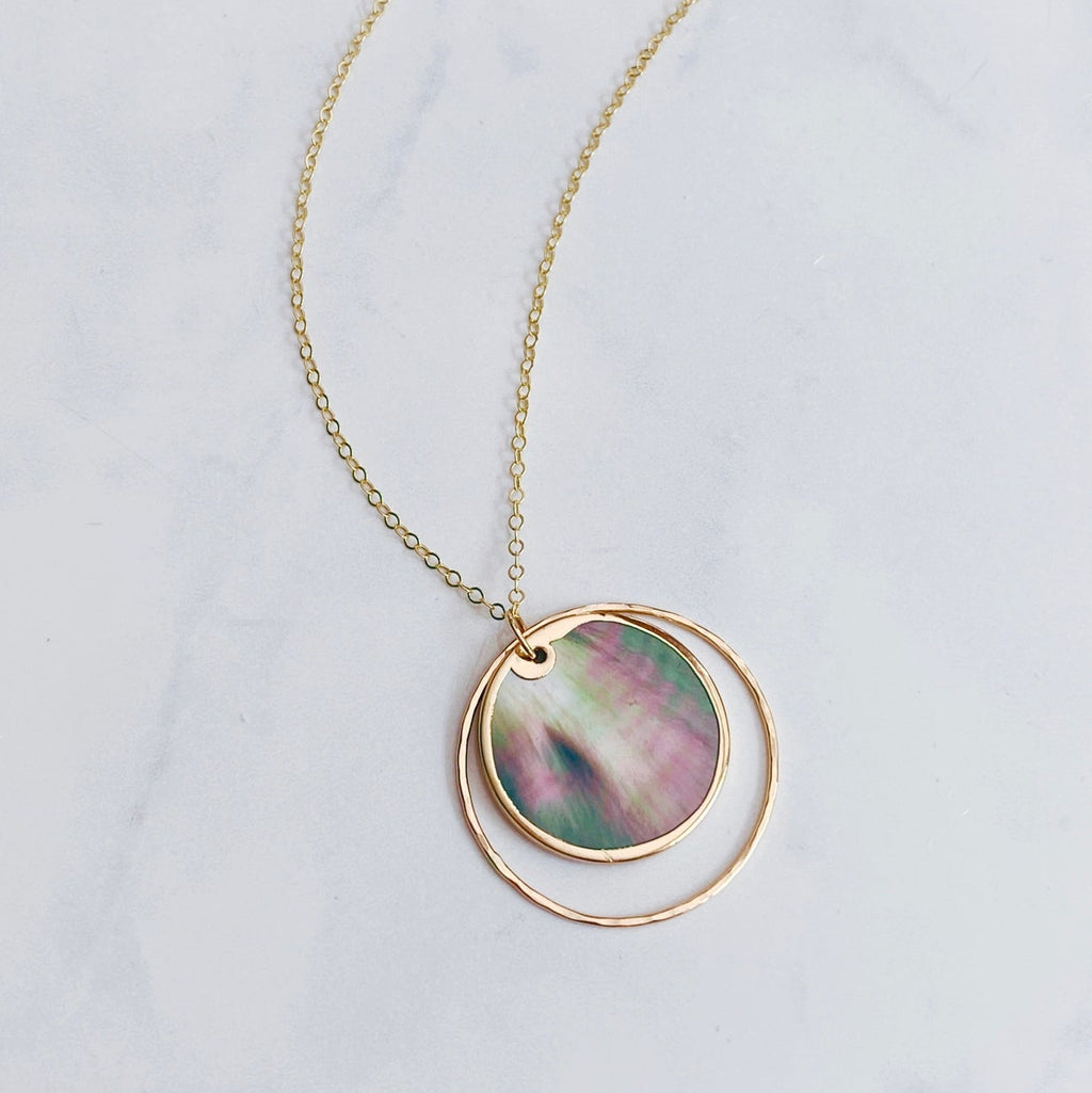 A gold chain necklace featuring a double-ring pendant, with the inner ring displaying a round iridescent shell inlay of pink, green, and blue tones, set against a light gray marble background.