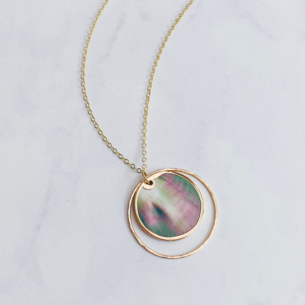 A gold chain necklace featuring a double-ring pendant, with the inner ring displaying a round iridescent shell inlay of pink, green, and blue tones, set against a light gray marble background.