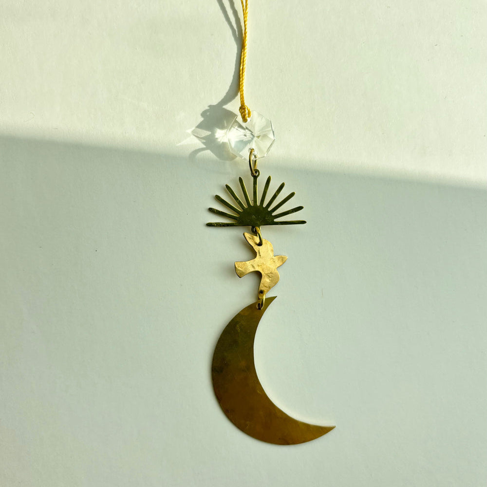 A brass crescent moon and sun pendant ornament with a bird charm, suspended from a gold cord with a clear faceted crystal bead, shown against a plain off-white background.
