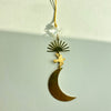 A brass crescent moon and sun pendant ornament with a bird charm, suspended from a gold cord with a clear faceted crystal bead, shown against a plain off-white background.