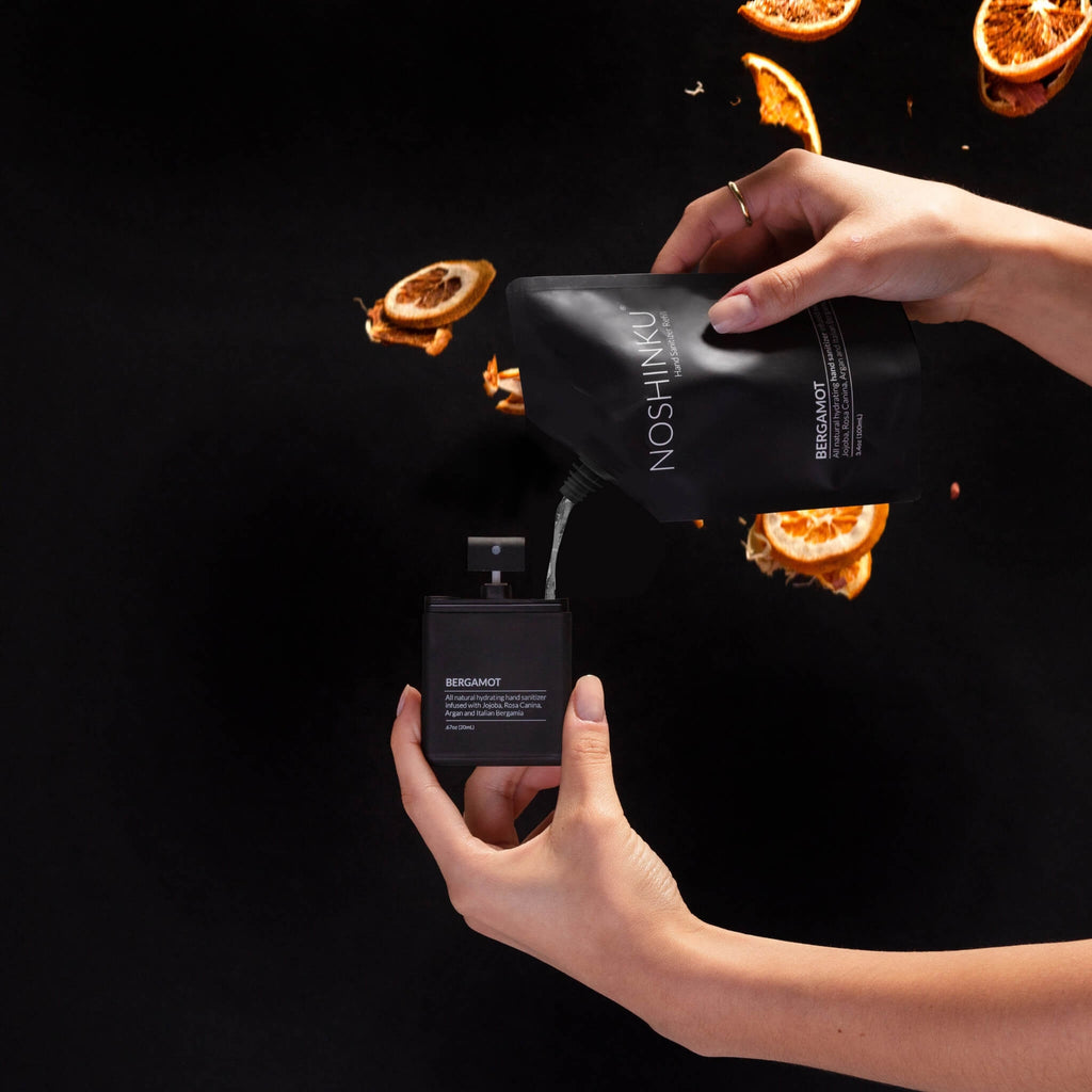 Person holding a black Noshkinu refill bottle and packaging against a dark background with orange slices.