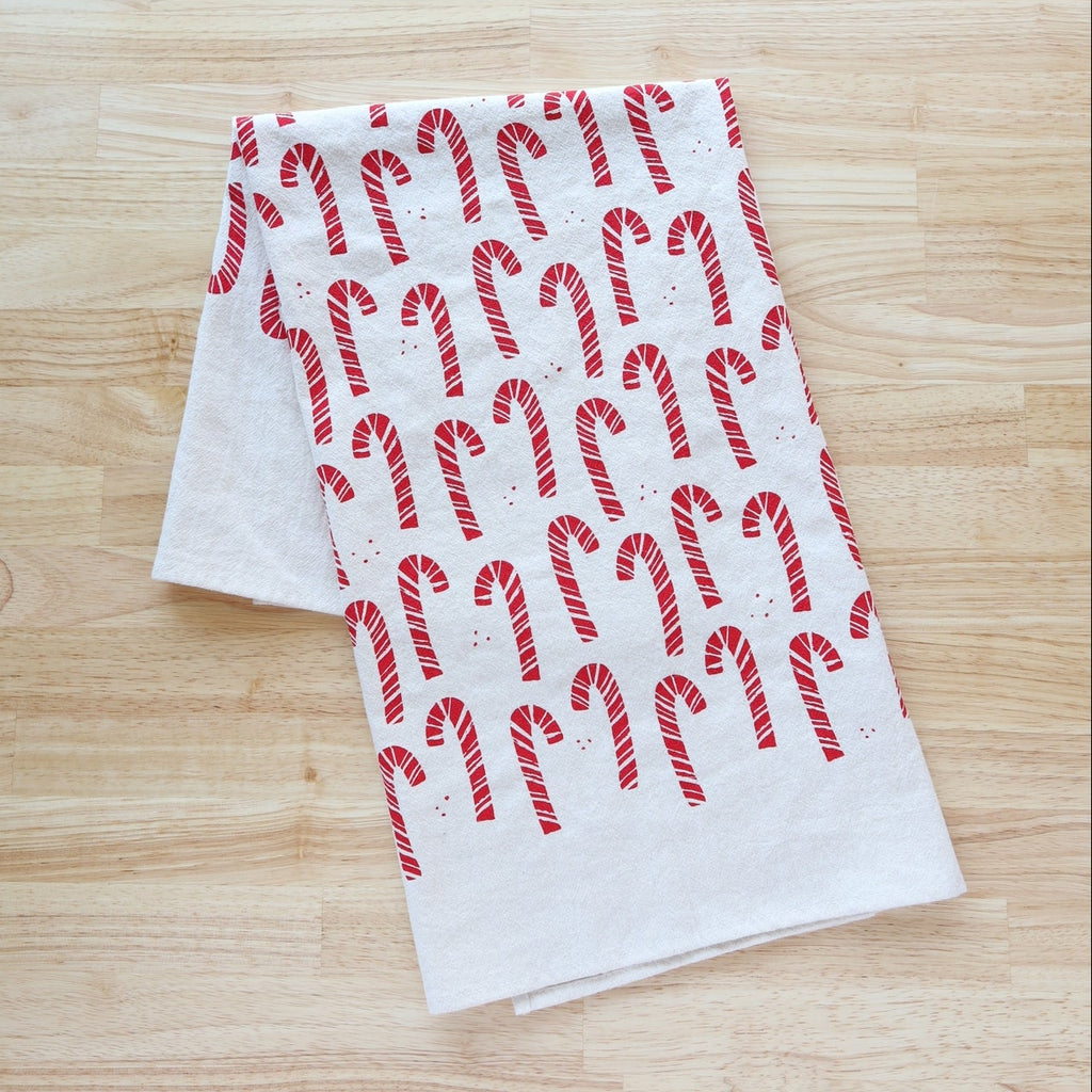 A white fabric tea towel with a red candy cane pattern, folded on a light wood grain surface.