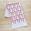 A white fabric tea towel with a red candy cane pattern, folded on a light wood grain surface.