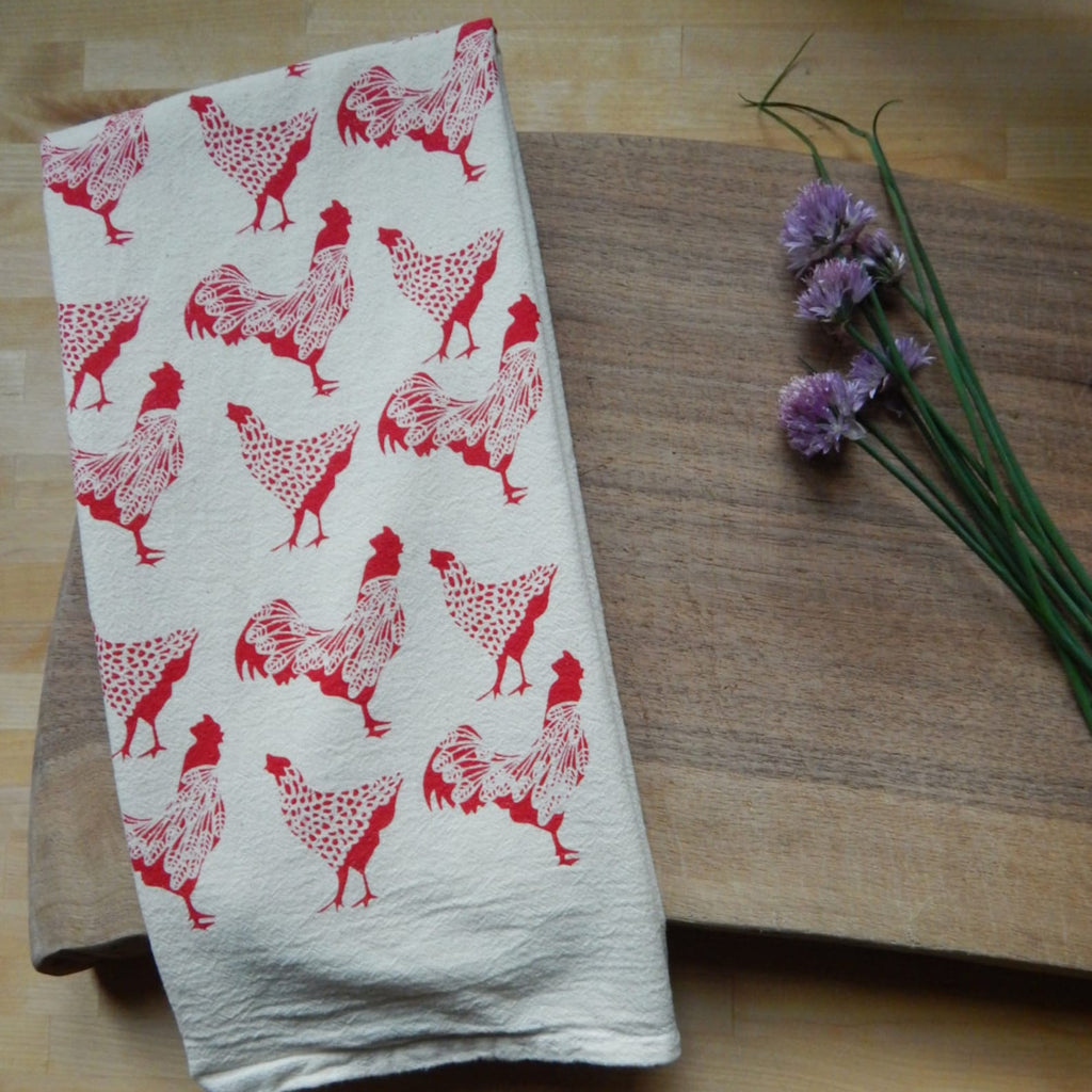White towel with red rooster pattern on a wooden surface with flowers.