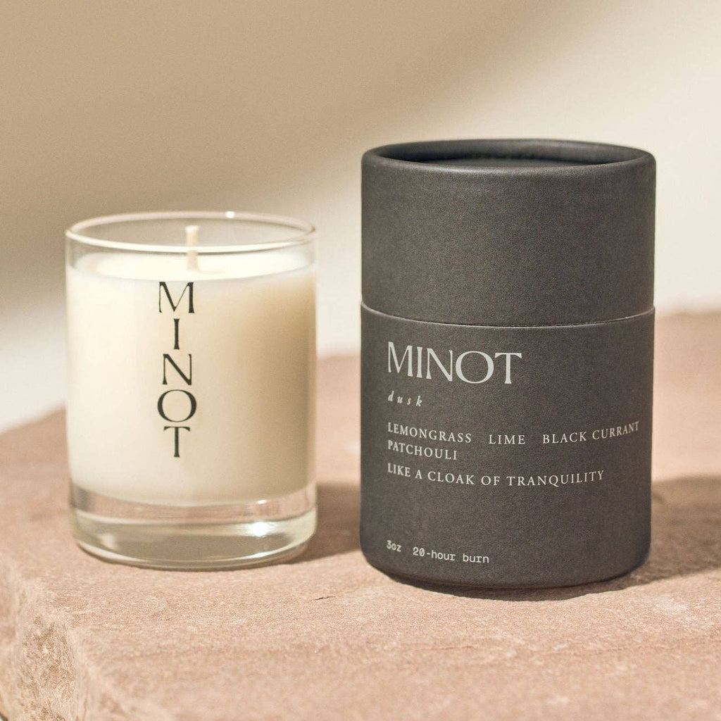 Two candles, one glass and one in black packaging, with 'MINOT' branding on a beige surface.