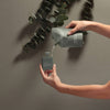 Person pouring liquid from a gray bottle labeled 'Noshinku' into another container against a neutral background with greenery.