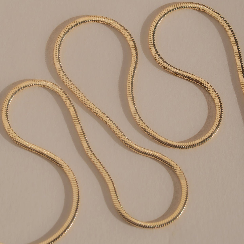A gold snake chain necklace draped in an S-shaped loop on a neutral beige background.