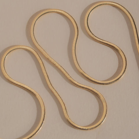A gold snake chain necklace draped in an S-shaped loop on a neutral beige background.