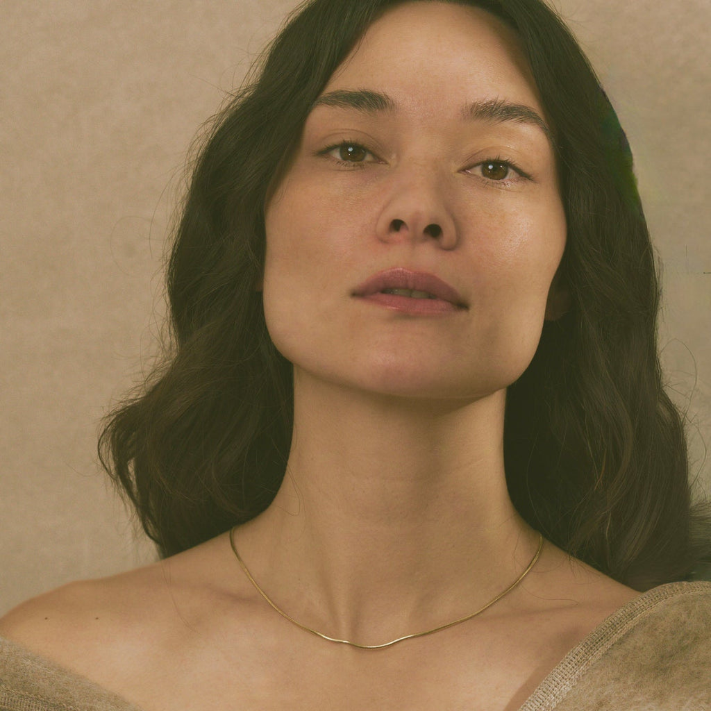 A close-up portrait of a woman with dark hair wearing a delicate gold snake chain necklace and a beige knit sweater against a neutral beige background.