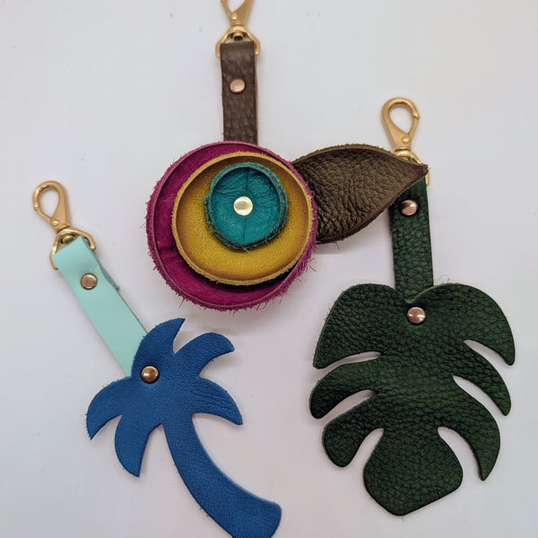 Variety of differently shaped bag charms: a blue palm tree, a magenta, yellow, and cyan blue flower, and a dark green monstera leaf.