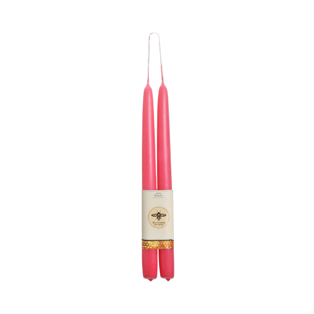 A pair of tall, slender pink taper candles standing upright side by side, wrapped together with a cream paper band featuring a bee logo and a gold glitter accent, isolated on a white background.