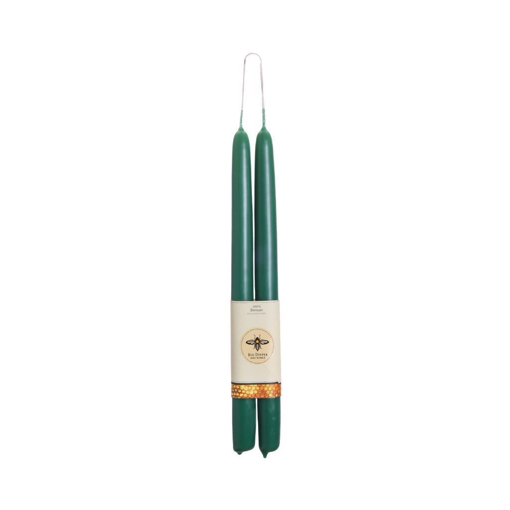 Two dark green taper candles wrapped together with a cream paper band featuring a bee logo and a gold patterned accent, set against a plain white background.