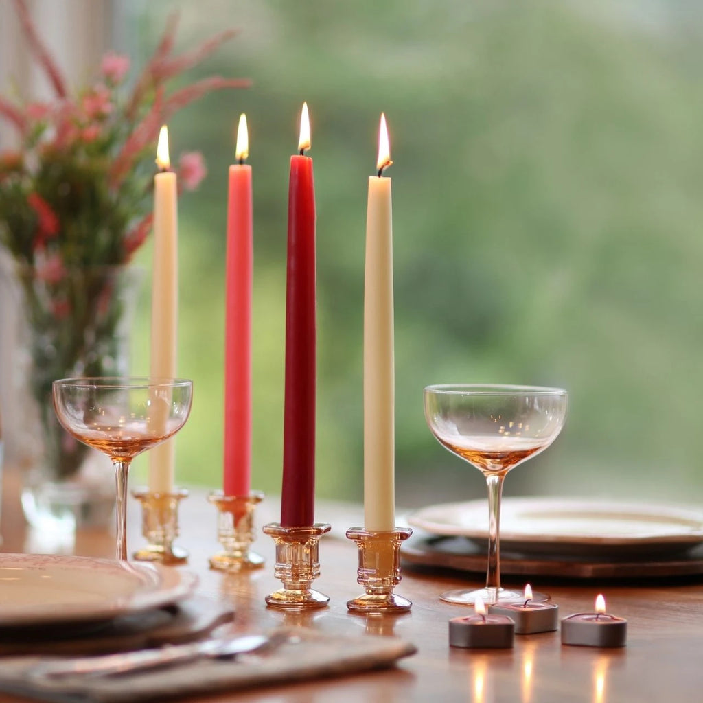 Four lit taper candles in glass holders arranged on a dining table, with wine glasses, plates, and small tealight candles in a softly blurred indoor setting.