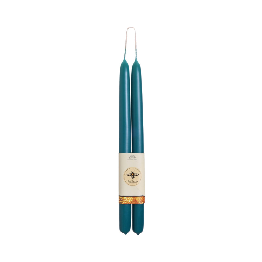 A pair of teal taper candles with a cream paper band label featuring a bee logo and a gold patterned accent, set against a plain white background.