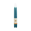 A pair of teal taper candles with a cream paper band label featuring a bee logo and a gold patterned accent, set against a plain white background.