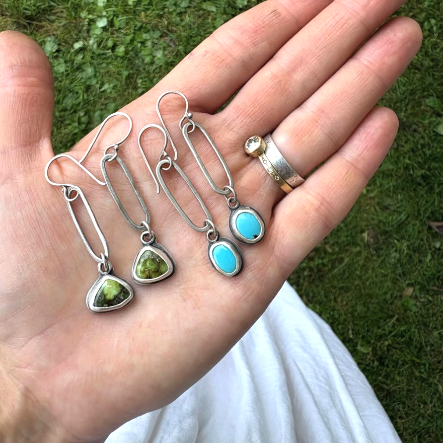 Hand holding four silver earrings with gemstones against a grassy background