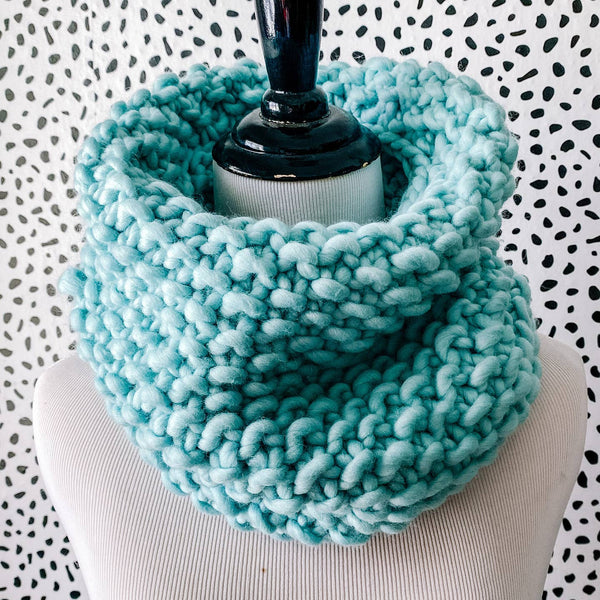 Light turquoise knitted cowl on a mannequin against a patterned background
