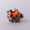 Small mechanical object with a red face, gold accents, and a mustache on a light gray background