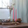 Two pink candles in gold holders on a wooden surface with a vase of flowers and a bowl in the background.