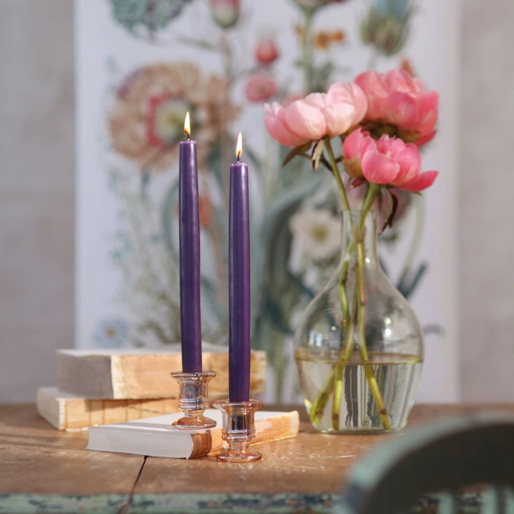 Two lit purple taper candles in clear glass holders on a rustic wooden table, next to a glass vase with pink flowers, with a blurred floral painting in the background.