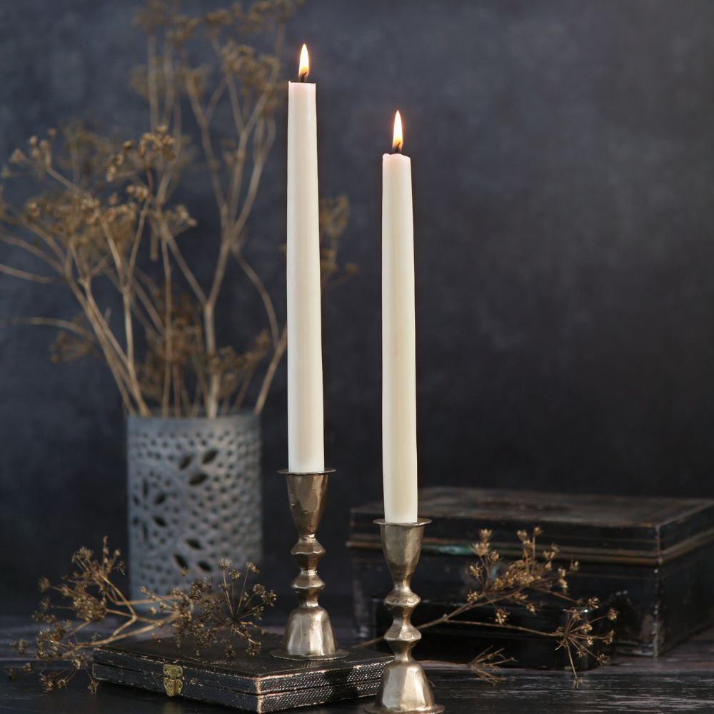 Two lit taper candles in silver-toned metal candlesticks, styled on a dark tabletop with dried flowers and a vase against a dark blue-gray background.