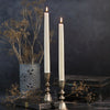 Two lit taper candles in silver-toned metal candlesticks, styled on a dark tabletop with dried flowers and a vase against a dark blue-gray background.