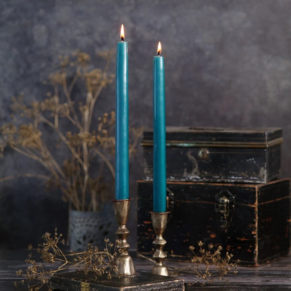 Two lit teal taper candles in ornate silver candlesticks, styled on a rustic wooden surface with dried flowers and stacked vintage black boxes against a dark textured background.