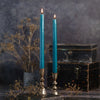 Two lit teal taper candles in ornate silver candlesticks, styled on a rustic wooden surface with dried flowers and stacked vintage black boxes against a dark textured background.