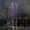 Two lit purple taper candles in ornate silver candlesticks, styled on a dark rustic surface with dried flowers and a vintage black chest against a dark textured background.