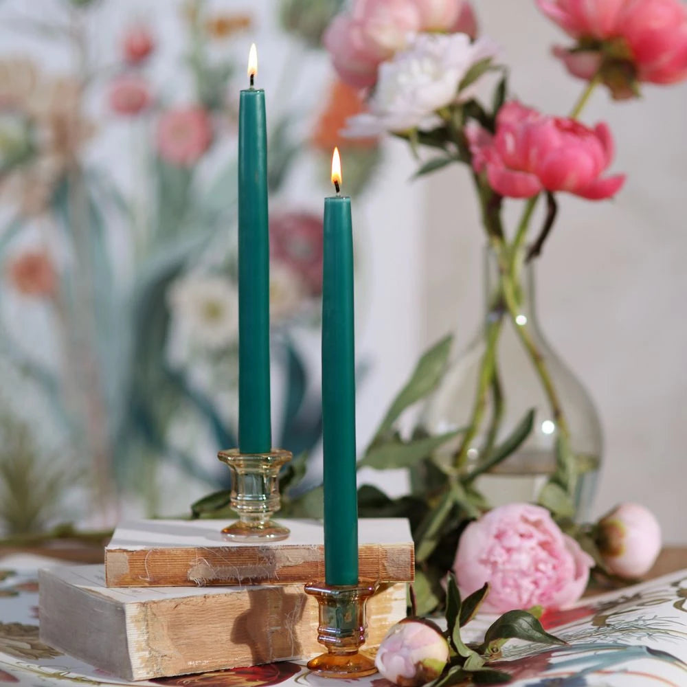 Two teal candles in wooden candle holders on a table with flowers in the background.