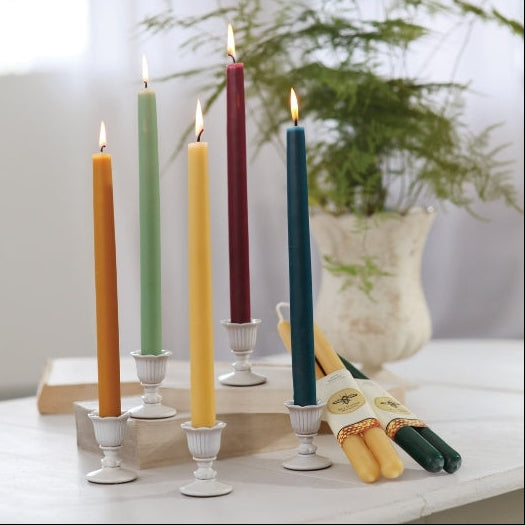 Four lit taper candles in different colors—orange, light green, mustard yellow, and dark blue—standing in white candle holders on a tabletop. A wrapped set of additional candles lies beside them, with a softly blurred indoor background and greenery in the distance.