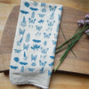A folded cream-colored linen tea towel with a blue botanical and butterfly pattern, styled on a wooden cutting board next to purple chive flowers.