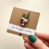 Hand holding a small bouquet of flowers on a kraft paper card with 'You Brighten My Day' text.