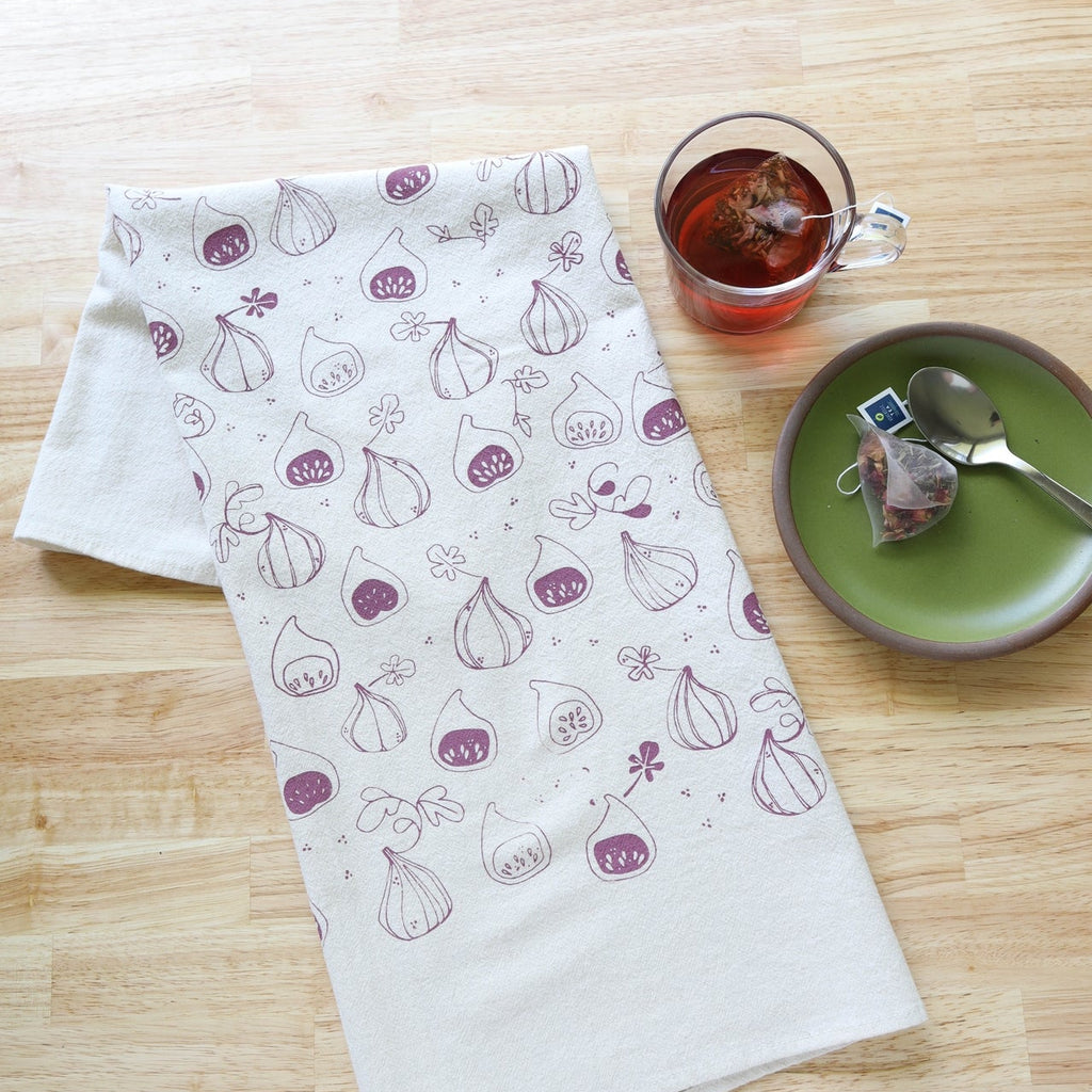Tea towel with fig pattern on a wooden surface with tea and a plate.