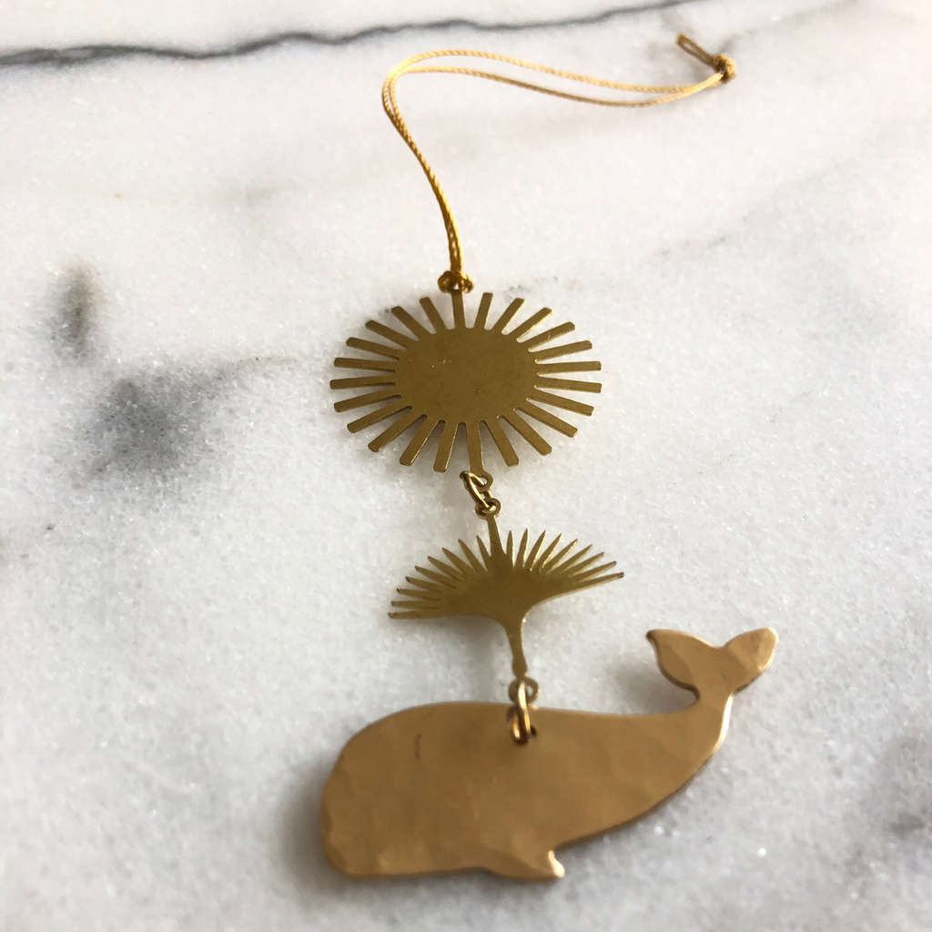 A gold-toned metal wall hanging ornament with a sunburst disc, a small fan-like piece, and a whale silhouette charm, suspended by a cord on a light marble surface.