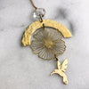 Brass pendant ornament featuring a hammered crescent moon, a cutout flower, and a small hummingbird charm, with a clear faceted bead at the top, photographed on a light marble-like surface.