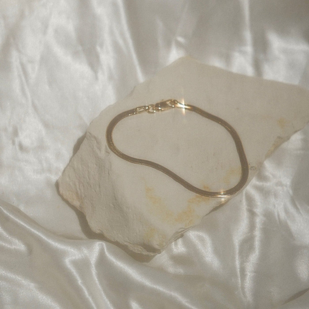 A delicate gold snake chain bracelet resting on a textured, irregularly shaped beige stone slab, set against a draped white satin fabric background.