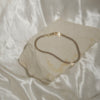A delicate gold snake chain bracelet resting on a textured, irregularly shaped beige stone slab, set against a draped white satin fabric background.